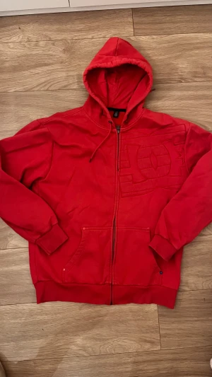 Röd DC hoodie med dragkedja - Riktigt fet röd hoodie från DC med stor broderad logga på bröstet. Tröjan har huva med snörning, dragkedja hela vägen och två fickor framtill. Perfekt för dig som gillar streetwear.