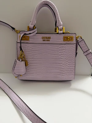 Ljuslila handväska från Guess - Snygg ljuslila handväska från Guess med croco-mönster och guldfärgade detaljer. Väskan har både handtag och axelrem, samt dekorativt hänglås och nyckel. Insidan är ljus med flera fack och dragkedja upptill. Perfekt för dig som gillar stilrena accessoarer med en twist. Använd en gång 