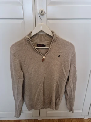 Beige stickad tröja Morris half zip - Snygg beige stickad tröja från Morris med half zip och ribbad krage. Liten svart broderad logga på bröstet och diskreta detaljer. Perfekt för dig som gillar klassisk stil med modern touch. Passar dig som vill ha en tröja med lite extra edge.