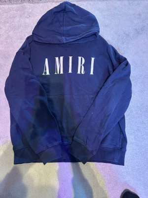 Marinblå AMIRI hoodie - Snygg marinblå hoodie från AMIRI med stort vitt logotryck på bröstet. Tröjan har huva, ribbade muddar och en klassisk känguruficka framtill. Perfekt för dig som gillar streetwear och vill ha en stilren look. Den har dock lite för liten huva och lite defekt på taggen men annars är den i bra skick skulle jag säga. Den passar ungefär xs-s men mer åt xs sidan. Skriv bara om ni har några funderingar!