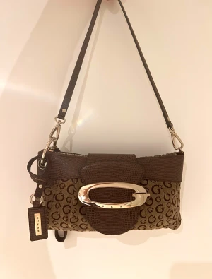 Brun axelväska från Guess med G-mönster - Snygg brun axelväska från Guess med klassiskt G-mönster över hela väskan. Väskan har detaljer i mörkbrunt skinnliknande material, silverfärgat spänne framtill och dragkedja upptill. Insidan har fack och en mindre ficka med dragkedja. Perfekt storlek för det viktigaste.