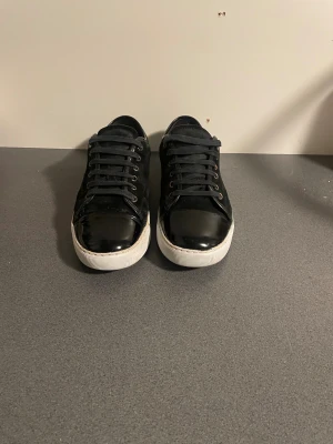 Svarta lanvins  - Säljer ett par svarta sneakers med snygg lackad tå och vit platt sula. Skorna har snörning och en mix av mocka och blankt material framtill, vilket ger en cool kontrast. Perfekta för dig som gillar stilrena och moderna skor.