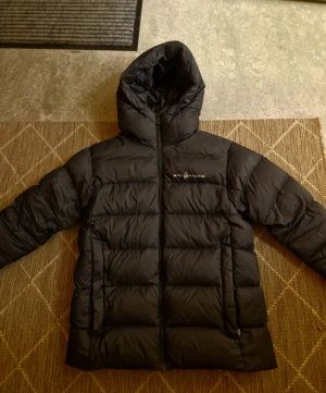 Svart Sail Racing dunjacka med huva - Svart dunjacka från Sail Racing med fyllning av Allied Feather + Down (600 fill power). Jackan har en stor huva, quiltad design och dragkedja framtill. Perfekt för kalla dagar och har en clean look med logga på bröstet.