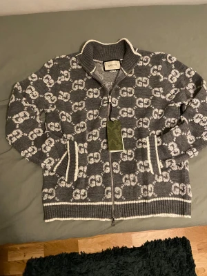 Grå stickad kofta från Gucci - Snygg grå stickad kofta från Gucci med vit GG-logga över hela plagget. Koftan har ribbad krage, muddar och nederkant, samt en silverfärgad dragkedja med Gucci-gravyr. Fina vita detaljer vid fickor och kanter. Perfekt för dig som gillar lyxiga märken.