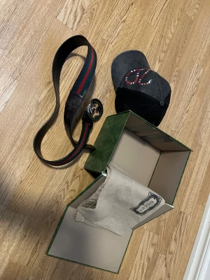 Gucci gott o blandat låda - Gucci bälte i svart skinn med klassiskt GG-spänne i metall. Bältet har ett randigt band i grönt och rött längs mitten, vilket ger en ikonisk Gucci-look. Levereras med originalförpackning och dustbag. Perfekt accessoar för att lyfta din outfit.