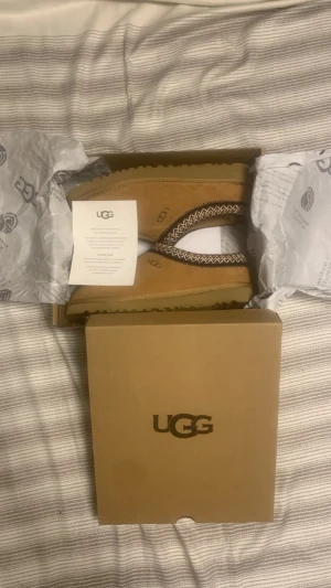 UGG bruna mocka slip-ins med mönster - Säljer ett par UGG slip-in skor i brun mocka med rund tå och platt sula. Skorna har ett snyggt zigzag-mönster runt öppningen och är fodrade med mjuk, vit päls på insidan för extra komfort. Perfekta för kalla dagar och chill hemma.