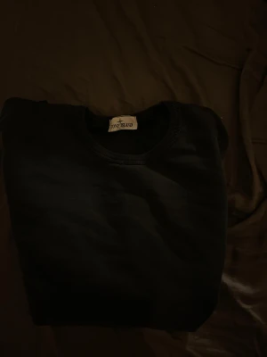 Svart sweatshirt från Stone Island - Svart crewneck sweatshirt från Stone Island med klassisk rund hals och ikonisk logotyp-patch på vänster ärm. Tröjan är tillverkad i mjuk bomull och har en clean look utan mönster. Perfekt för dig som gillar streetwear och vill ha något stilrent.