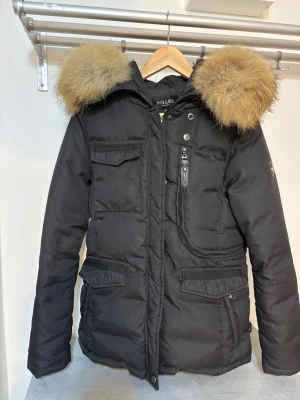 Hollies jacka  - Säljer en svart dunjacka från Hollies med stor fluffig päls runt huvan. Jackan har flera fickor med lock, dragkedja och knappar framtill samt snygga detaljer. Perfekt för kalla dagar och riktigt varm. Klassisk modell med lång ärm och markerad midja. Hollies livingo dam dunjacka med äkta päls. 