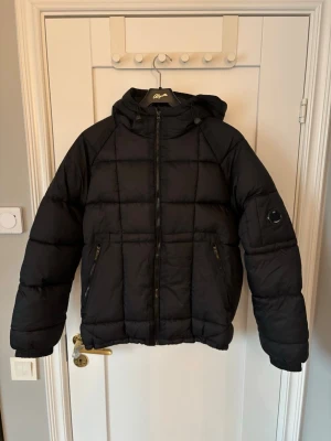 Svart pufferjacka från C.P. Company - Svart pufferjacka från C.P. Company med huva och dragkedja framtill. Jackan har två snedställda fickor med dragkedja och en ikonisk linsdetalj på ärmen. Säljs då den inte längre kommer till användning. Passar bra som tunn vinterjacka men ännu bättre till våren. 9/10 skick