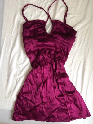 Vinröd satin klänning med cut-out - Vinröd kort klänning i satin med smala axelband och snygg cut-out detalj framtill. Klänningen har en figurnära passform och en lätt glansig yta som ger en lyxig känsla. Perfekt för dig som vill sticka ut med en trendig look.