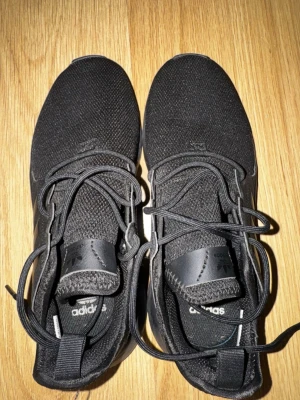 Svarta Adidas sneakers med snörning - Snygga helsvarta Adidas sneakers med klassiska tre ränder på sidan. Ovandelen är i mesh-liknande syntetmaterial och skorna har snörning för bra passform. Perfekta för dig som gillar stilrena och sportiga skor.