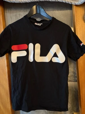 Svart FILA t-shirt med stor logga - Svart t-shirt från FILA med stor, ikonisk logga i vitt och rött på bröstet. Klassisk rund hals och korta ärmar. Perfekt för dig som gillar sportig och stilren streetwear. Tillverkad i mjuk bomull för skön känsla hela dagen.