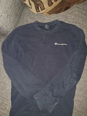 Mörkblå sweatshirt från Champion - Mörkblå sweatshirt från Champion med klassisk logga broderad i vitt på bröstet. Tröjan har rund halsringning, långa ärmar och ribbade muddar vid ärmslut och nederkant. Perfekt för en avslappnad och sportig stil.