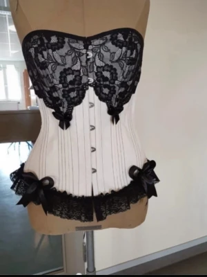 Vit och svart spetskorsett med rosetter - Snygg korsett i vitt med svarta spetsdetaljer och dekorativa rosetter. Överdelen har blommig spets och knäppning fram, medan baksidan har snörning för justerbar passform. Svart spetskant nedtill och fina banddetaljer ger en elegant look.(hem sydd) diskuterar priset vid snabb affär