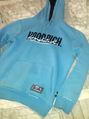 Blå hoodie från Hoodrich - Snygg ljusblå hoodie från Hoodrich med stor magficka och tryckt logga på bröstet. Huva med dragsko och ribbade muddar. Märkeslapp nedtill och coolt tryck även på ryggen. Perfekt för dig som gillar streetwear och vill sticka ut.