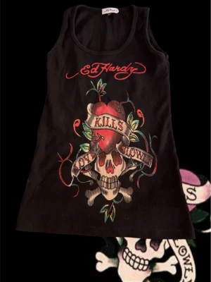 Svart Ed Hardy linnetopp med tryck - Svart linnetopp från Ed Hardy med ikoniskt dödskalle- och hjärtmotiv samt texten 'Love Kills Slowly' på framsidan. Toppen har ribbad struktur, ärmlös design och röd logga både fram och bak. Perfekt för dig som gillar edgy streetstyle.