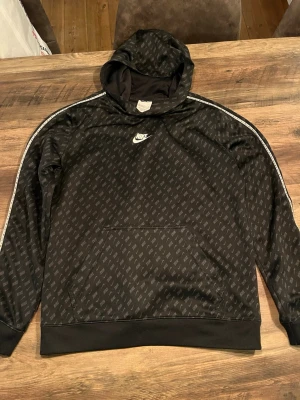 Svart Nike hoodie med logomönster - Snygg svart hoodie från Nike med all-over logomönster och vit Nike-logga på bröstet. Hoodien har huva, magficka och vita detaljer längs ärmarna. Tillverkad i ett mjukt och ventilerande material.