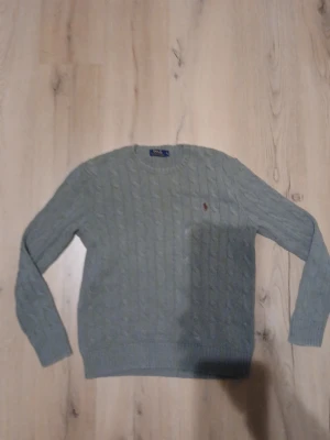 Ralph Lauren tröja - Snygg ljusgrön kabelstickad tröja från Polo Ralph Lauren med rund halsringning och brunröd broderad logga på bröstet. Tröjan har långa ärmar och ribbade muddar vid ärmslut och nederkant. Som du ser på bild 3 så har den två ställen där den är lite blekt. Perfekt för dig som är grisch! PS det står att den är grå för det ser ut som att den är det mest men är egentligen ljusgrön!