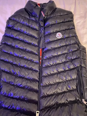 Svart dunväst från Moncler - Svart glansig dunväst från Moncler med klassisk logga på bröstet. Västen har dragkedja framtill och röd insida med Moncler-tryck. 