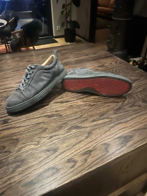 Louboutins skor grå  - Snygga grå Loubitins i mocka med klassisk låg profil och snörning. Skorna har en unik röd sula med mönsterdetalj och beige innerfoder. Perfekta för dig som gillar stilrena och tidlösa sneakers med en twist.