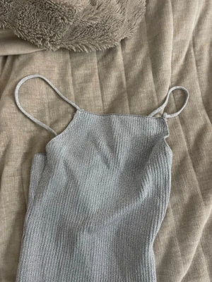 Grå ribbad crop top med tunna band - Säljer en stilren grå crop top med tunna axelband och ribbad struktur med lite glitter. Toppen har en rak halsringning och är gjord i ett mjukt, stretchigt material som sitter snyggt på kroppen. Perfekt att matcha med jeans eller kjol. Den har bara blivit till användning en gång. Perfekt fest topp! Storlek S från H&M