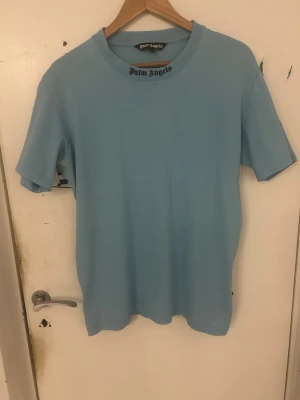 Ljusblå Palm Angels t-shirt - Säljer en ljusblå t-shirt från Palm Angels med logga tryckt i svart vid halsen. Klassisk rund halsringning och korta ärmar. Tillverkad i mjuk bomull för skön känsla och clean look. Perfekt för dig som gillar streetwear och enkel stil.