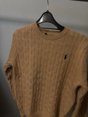 Beige kabelstickad tröja Polo Ralph Lauren - Säljer en beige kabelstickad tröja från Polo Ralph Lauren med rund halsringning och klassisk marinblå logga broderad på bröstet. Tröjan har ribbade muddar vid ärmslut och nederkant och är gjord i mjukt material. Perfekt för dig som gillar stilrena plagg.