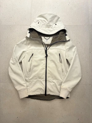  Windbreaker  C.P. Company - Stilren beige vindjacka från C.P. Company med svart dragkedja och fyra fickor framtill. Jackan har huva, långa ärmar och är tillverkad i ett slätt syntetmaterial. Perfekt för blåsiga dagar och enkel att matcha med olika outfits.           Den är i storlek S och fleece på insidan som håller dig varm. Vid frågor eller funderingar är det bara att skriva!!🤗🤗