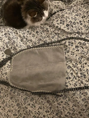 Grå mocka handväska med kedja - Snygg handväska i ljusgrå mocka med dekorativa kedjehandtag och kantdetaljer. Väskan har en mjuk, slät yta och en cool metallbricka vid handtaget. Perfekt för dig som gillar stilrena accessoarer med en twist.