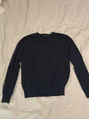 Mörkblå stickad tröja från Polo Ralph Lauren - Mörkblå stickad tröja från Polo Ralph Lauren med rund halsringning och diskret röd logga broderad på bröstet. Tröjan har långa ärmar och ribbade muddar vid ärmslut och nederkant. Perfekt för en stilren och klassisk look.