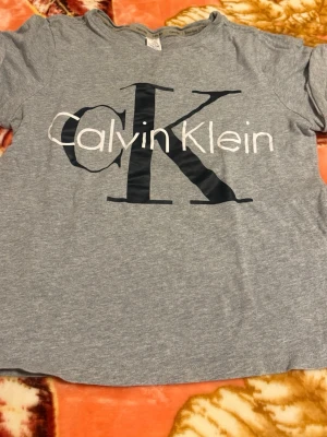 Grå Calvin Klein pyjamaströja - Mysig grå pyjamaströja från Calvin Klein med stort svart och vitt logotryck på bröstet. Tillverkad i mjukt material som passar perfekt för avslappnade kvällar hemma. Rund halsringning och korta ärmar ger en klassisk och bekväm look.