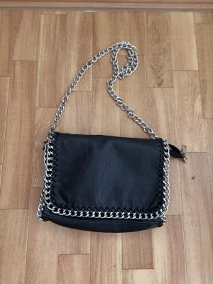 Tiamo väska - Snygg svart axelväska med  silverfärgad kedjerem och  kedja runt kanterna. Väskan har ett lock framtill och en dragkedjeficka på baksidan. 