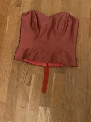 Rosa satin korsett med sweetheart-ringning - Trendig rosa korsett i satin med markerad sweetheart-ringning och avtagbart band framtill. Korsetten har dold dragkedja bak.