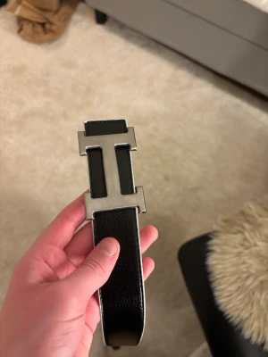 Vändbart läderbälte H-spänne - Snyggt vändbart bälte  med ikoniskt H-spänne i metall. Ena sidan är svart och den andra vit, båda i slätt läder. Perfekt accessoar för att lyfta din outfit med en lyxig touch och klassisk design.