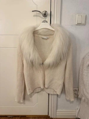 Beige cardigan jacka med päls - Ribbstickad beige cardigan jacka med stor, fluffig päls i vitt runt kragen. Jackan har långärmad design och en öppen front, perfekt för att lyfta din outfit med en lyxig touch. Storlek XL, sitter som S 