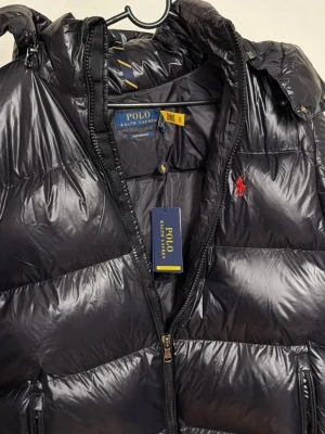 Polo Ralph Lauren svart dunjacka - Svart dunjacka från Polo Ralph Lauren med glansig finish, huva och dragkedja framtill. Jackan har Polo-logga broderad i rött på bröstet och flera praktiska fickor med dragkedja. Perfekt för kyliga dagar.