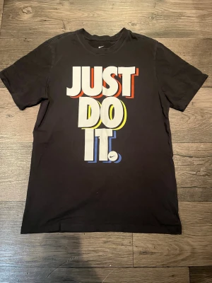 Svart Nike t-shirt med tryck - Svart t-shirt från Nike med stort färgglatt 'JUST DO IT.'-tryck på framsidan. Klassisk passform med rund halsringning och korta ärmar. Perfekt för en sportig och avslappnad stil.
