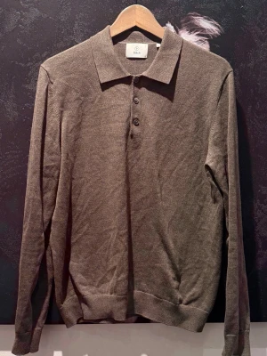 Brun långärmad pikétröja från Bläck  - Snygg brun långärmad pikétröja från Bläck med klassisk krage och tre knappar framtill. Tillverkad i mjukt, tunt material som ger en stilren look. Perfekt för dig som gillar en clean och enkel stil.