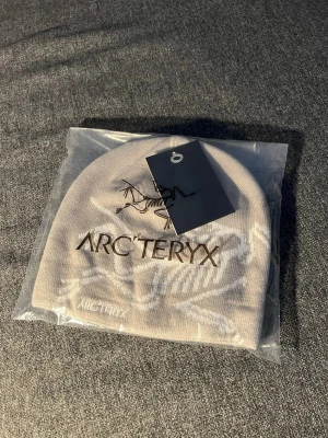 Grå  mössa - Snygg beige mössa  med broderad logga och fossilmotiv framtill. Tillverkad i mjukt stickat material som sitter skönt på huvudet. Perfekt för dig som vill ha en stilren och trendig accessoar till hösten och vintern.