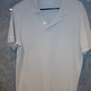 Beige pikétröja från Dressmann - Klassisk beige pikétröja från Dressmann med korta ärmar och krage. Tröjan har två knappar framtill och är tillverkad i ett mjukt bomullstyg som känns skönt mot huden. Perfekt för en clean och stilren look.