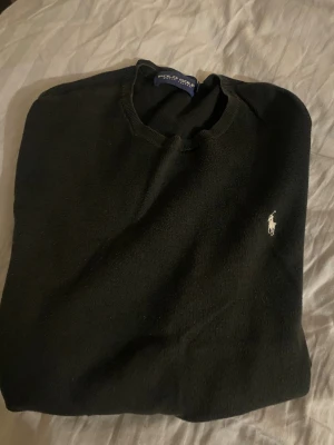 Svart tröja från Polo Ralph Lauren - Klassisk svart tröja från Polo Ralph Lauren med rund halsringning och den ikoniska vita logobroderingen på bröstet. Tröjan har ribbade muddar vid ärmslut och nederkant, perfekt för en stilren och enkel look.
