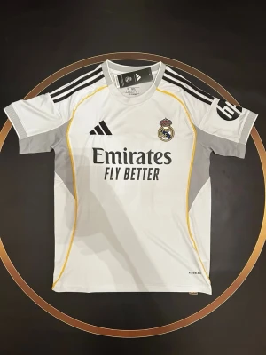 Real Madrid matchtröja Adidas vit - Snygg vit Real Madrid matchtröja från Adidas med svarta detaljer och gula linjer. Tröjan har klubbmärke på bröstet, Adidas-logga och sponsortryck 'Emirates Fly Better' framtill. Tillverkad i lätt och ventilerande material, perfekt för fotbollsträning. S och L