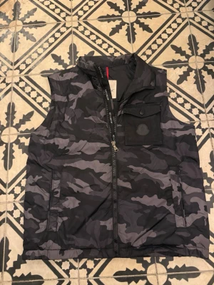 Camo dunväst från Moncler - Snygg dunväst från Moncler i svart och grått camomönster. Västen har hög krage, dragkedja framtill, två sidofickor och en bröstficka med knapp. Perfekt för lager-på-lager och streetwear-stil.