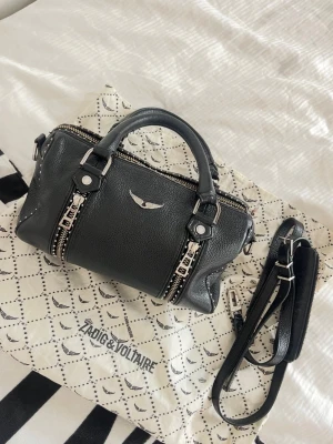 Svart Zadig & Voltaire handväska - Säljer en svart sunny bag från Zadig & Voltaire i skinn med silverfärgade detaljer och dubbla dragkedjor framtill. Väskan har både handtag och avtagbar axelrem, samt den ikoniska loggan i metall på framsidan. Perfekt storlek för det viktigaste. Köpt för 5000kr och är ganska andvänd, men ändå i ett väldigt bra skick. Kommer även med Zadig & Voltaire påsen som visas på bilden. Detta har varit min älsklings väska länge och jag lovar er man får plats med ALLT I DEN!! 