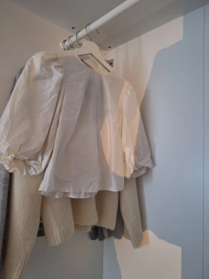 Vit blus med puffärmar och rosett - Vit blus med stora puffärmar och svart rosett i ryggen. Blusen har en rak passform och är tillverkad i ett lätt tyg som ger en luftig känsla. Perfekt för dig som gillar trendiga detaljer och en feminin look.