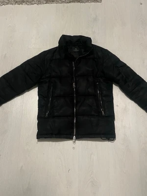Svart pufferjacka med dragkedjor - Svart pufferjacka från Bruuns bazaar med hög krage och två snedställda fickor med dragkedjor framtill. Jackan har quiltad design och en extra ficka med knapp på ena ärmen. Perfekt för kalla dagar och enkel att matcha. Luvan är tyvärr borta 