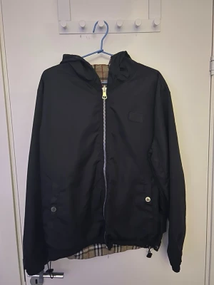 Burberry Windbreaker  - Snygg svart vindjacka med huva och dragkedja framtill. Jackan har ett klassiskt rutigt foder och justerbar nederkant med snörning. Perfekt för blåsiga dagar och enkel att matcha till olika outfits. Plagget kan vändas ut och in så de blir olika mönster, använd 2 gånger.