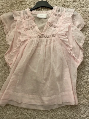 Ljusrosa volangblus från Neo Noir - Supersöt ljusrosa blus från Neo Noir med volangdetaljer och v-ringning. Blusen är tillverkad i ett tunt, luftigt tyg och har romantiska spetsdetaljer samt korta ärmar med volanger. Perfekt för en feminin och trendig look.