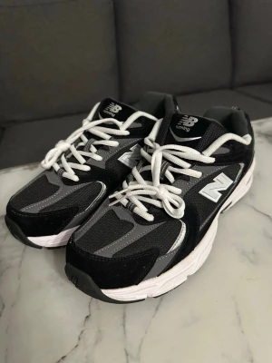 New Balance svarta 530 - Snygga New Balance sneakers i svart och grått med vita detaljer och klassisk snörning. Skorna har en sportig design med mesh och syntetmaterial, samt den ikoniska N-loggan på sidan. Perfekta för dig som gillar streetwear och bekväma skor.