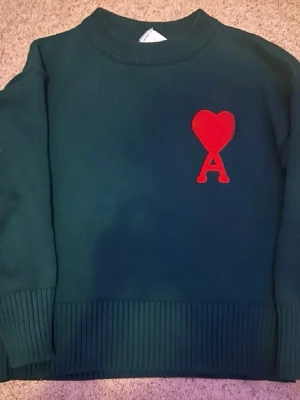 Grön stickad tröja från Ami Paris - Mörkgrön stickad tröja från Ami Paris med röd hjärtlogga och bokstaven A på bröstet. Tröjan har rund halsringning och breda ribbade muddar vid ärmslut och nederkant. Perfekt för dig som gillar stilrena detaljer och streetwear.